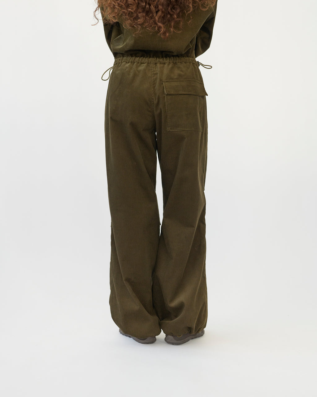The model is wearing a pair of wide-leg corduroy trousers in olive with an adjustable elastic waistband. White background, back view.